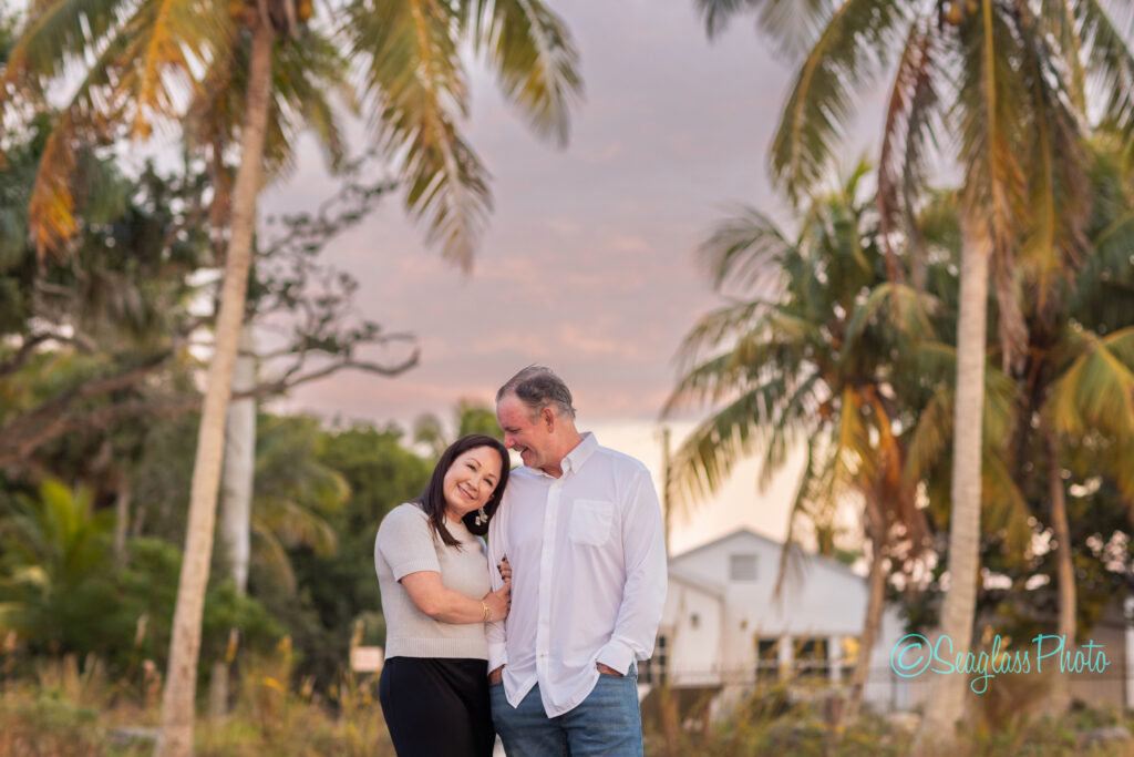 A smiling couple stands close together under palm trees at sunset, with a white house and lush greenery in the background. The woman rests her head on the mans shoulder, and both look relaxed and happy. Vero Beach Photoshoot