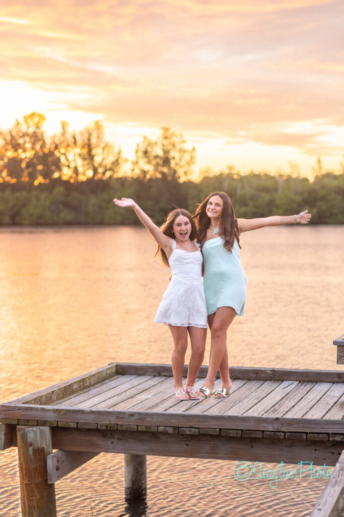 Two girls in summer dresses stand on a wooden dock by the water at sunset, smiling with their arms raised. The background shows trees and an orange sky reflected on the calm water. Vero Beach Photoshoot