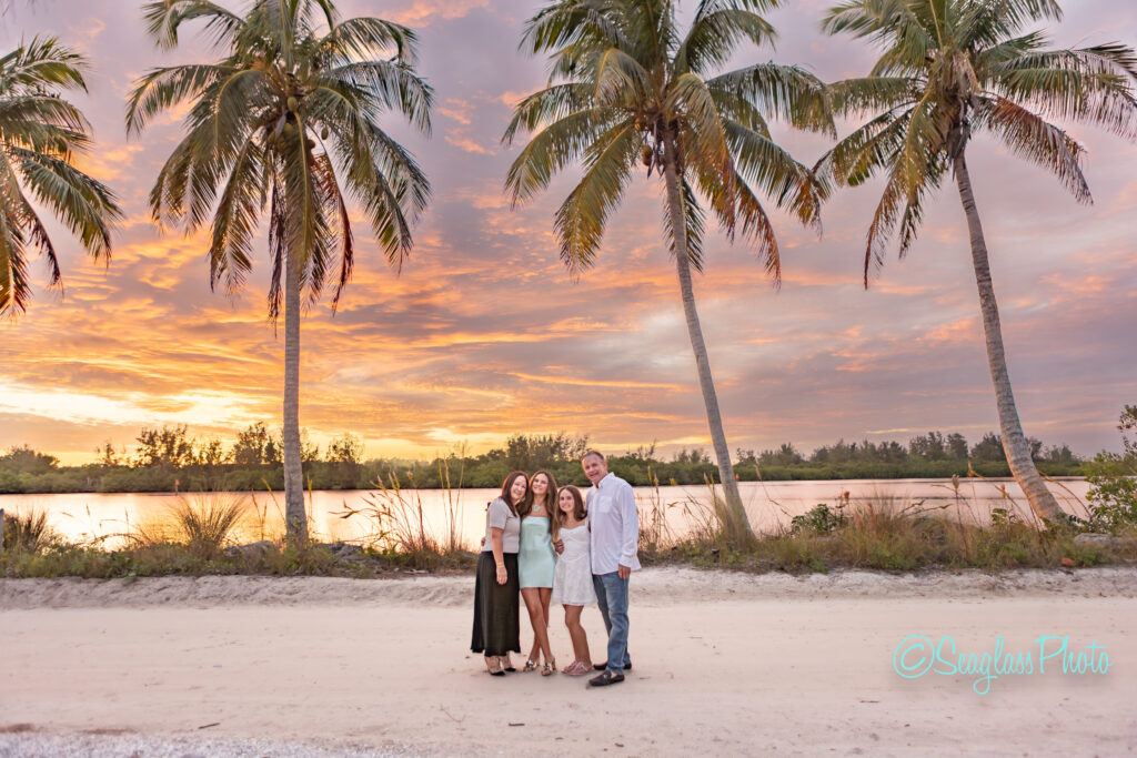 Four people stand closely together, smiling beneath tall palm trees at sunset. The sky is vibrant with orange and purple hues, and a calm body of water reflects the colors in the background. Vero Beach Photoshoot