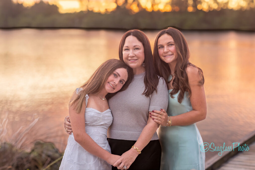 A woman stands between two teen girls, all smiling and hugging by a lake at sunset. The girls wear light dresses, and the woman wears a gray top and black skirt. The water and trees are softly lit in the background. Vero Beach Photoshoot