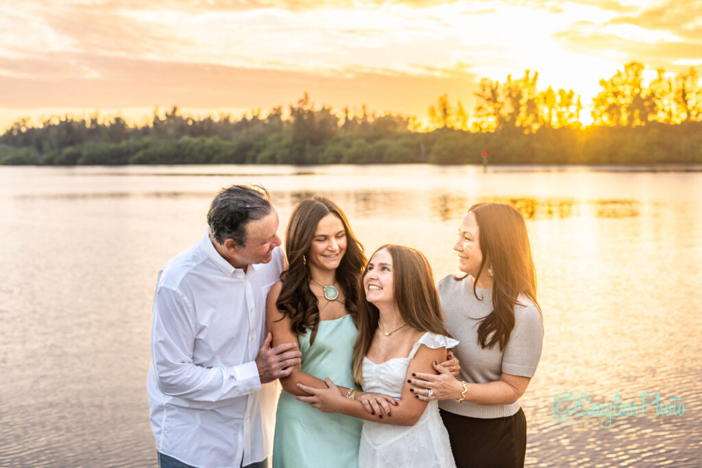 A family of four stands together by a lake at sunset, smiling and embracing. The two parents stand on either side of their two daughters, all dressed in light-colored clothes, with trees and a golden sky in the background. Vero Beach Photoshoot