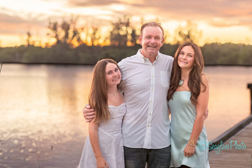 A man stands between two young women by a lake at sunset, all smiling. They are dressed in light-colored, summery clothes, with trees and a glowing sky in the background. Vero Beach Photoshoot