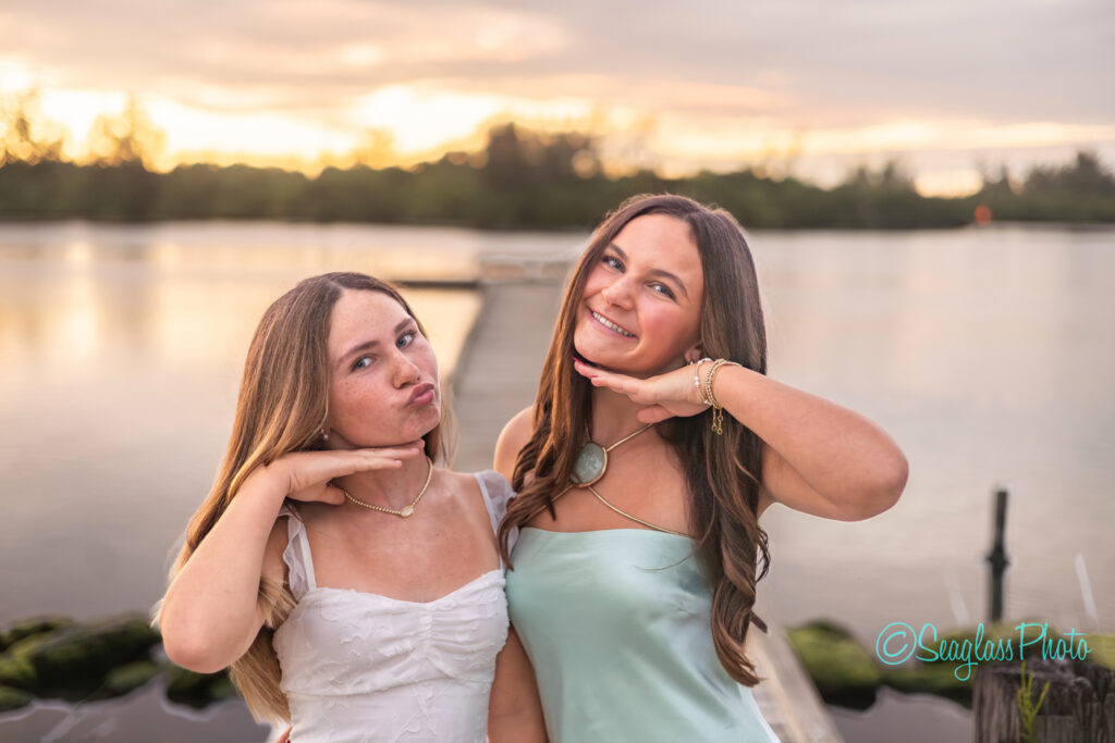 Two young women pose playfully with their hands under their chins, standing on a dock by a calm lake at sunset. They wear light-colored dresses and smile, with trees and soft sunlight in the background. Vero Beach Photoshoot
