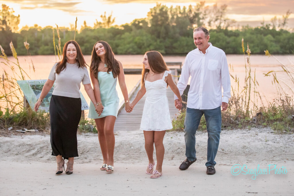A family of four, two adults and two girls, walk hand in hand on a sandy path by the water at sunset, smiling and dressed in light, summery clothes. Trees and a wooden dock are visible in the background. Vero Beach Photoshoot