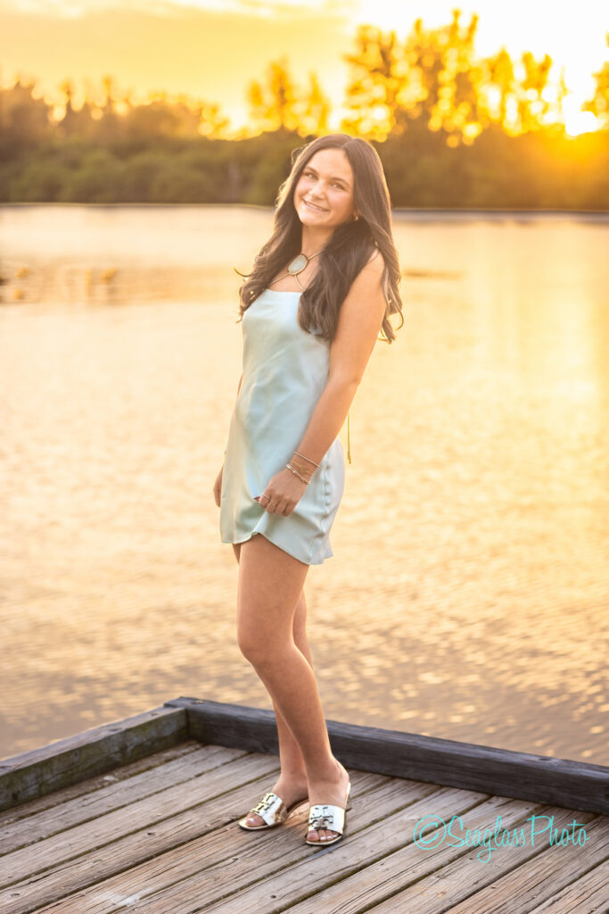 A young woman in a light blue dress stands barefoot on a wooden dock by a lake at sunset, smiling with trees and golden light reflected in the water behind her. Vero Beach Photoshoot