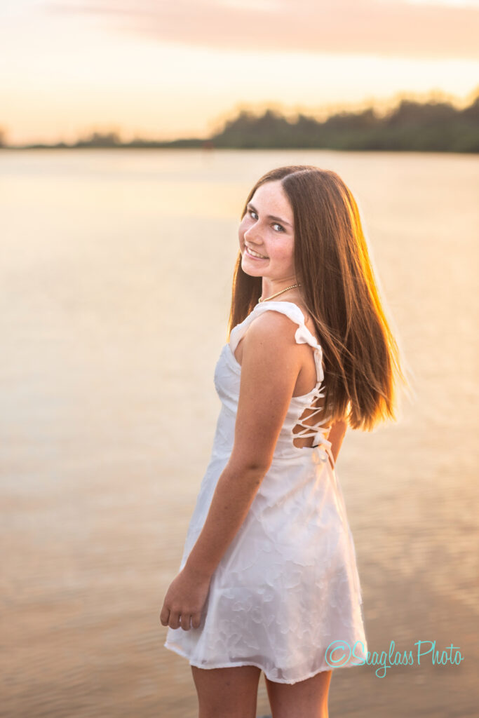 A teenage girl with long brown hair, wearing a white dress, stands by a calm lake at sunset, smiling and looking back over her shoulder. Vero Beach Photoshoot