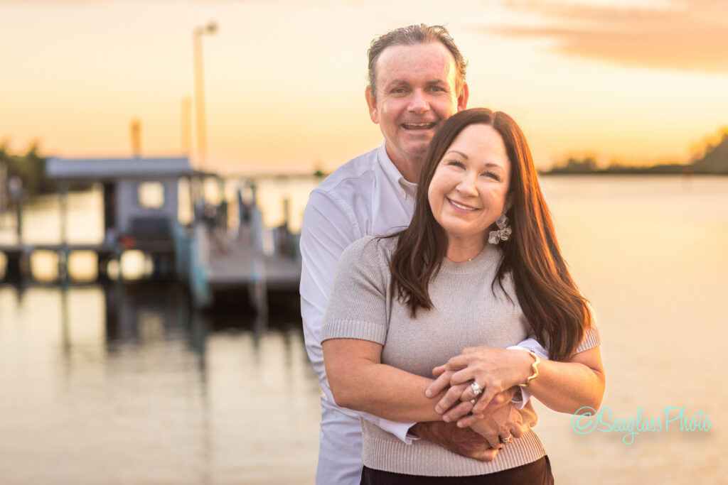 A smiling couple embraces by the water at sunset, with a dock and soft orange sky in the background. The man stands behind the woman, both looking happy and relaxed. Vero Beach Photoshoot