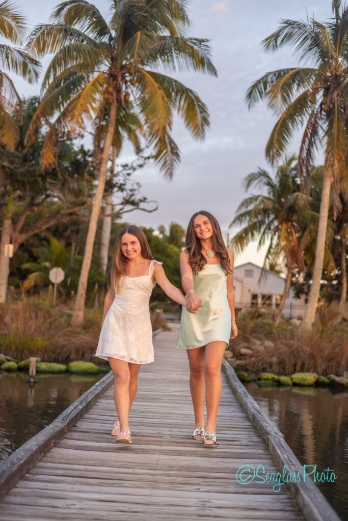 Two young women in light dresses walk hand-in-hand along a wooden dock over water, palm trees and a house in the background, with warm evening light illuminating the scene. Vero Beach Photoshoot