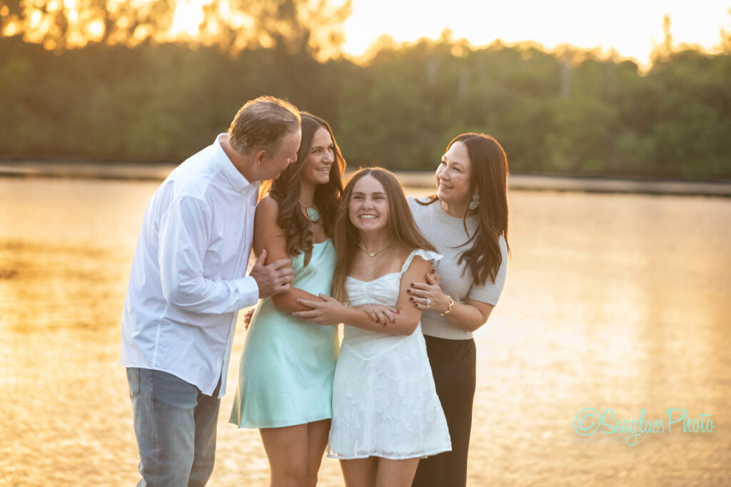 A happy family of four stands by the water at sunset, smiling and laughing together. The two girls wear light dresses, and the parents stand on either side, all enjoying a warm, golden moment outdoors. Vero Beach Photoshoot