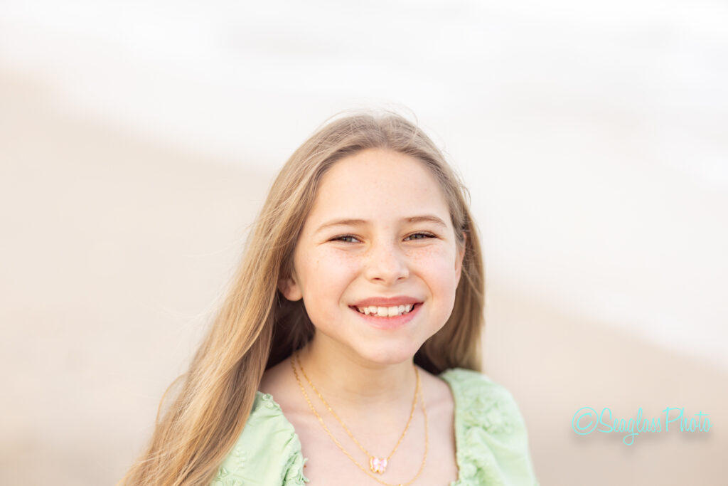 A young girl with long blonde hair smiles at the camera. She is wearing a light green top and a gold necklace. The background is softly blurred, suggesting a beach setting. Vero Beach Photoshoot