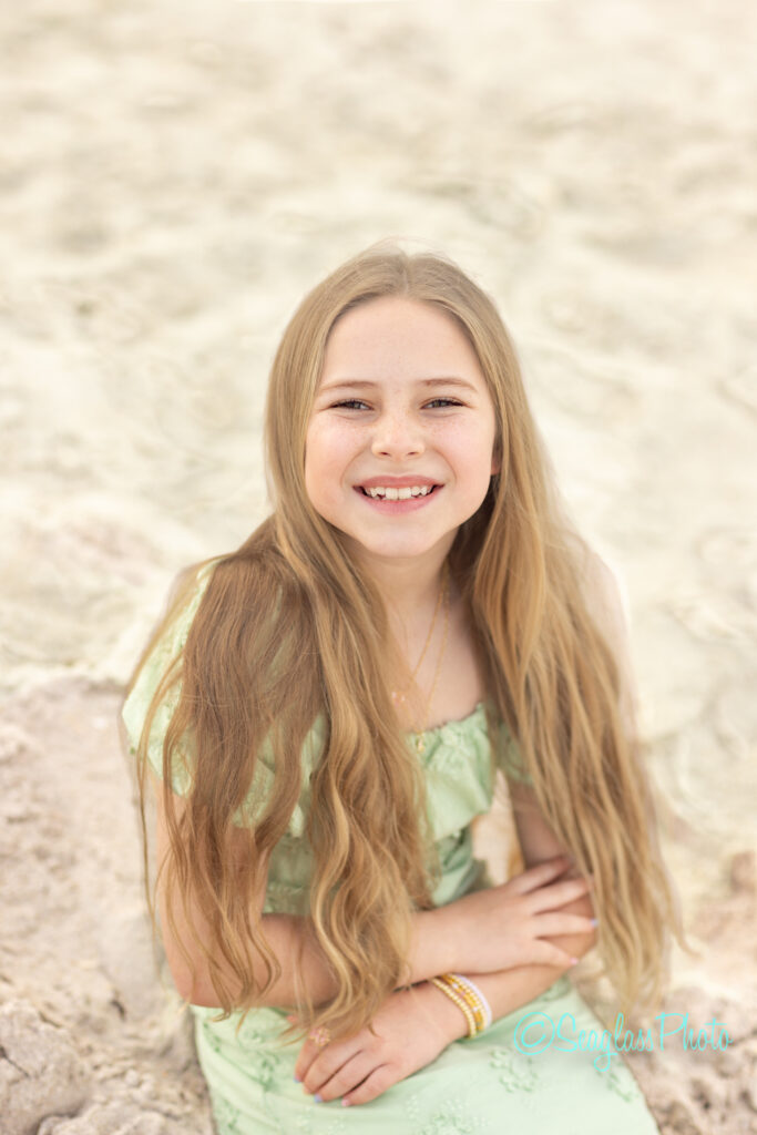A young girl with long blonde hair, wearing a light green dress and bracelets, sits on sand and smiles at the camera. The background is blurred, showing more sandy beach. Vero Beach Photoshoot