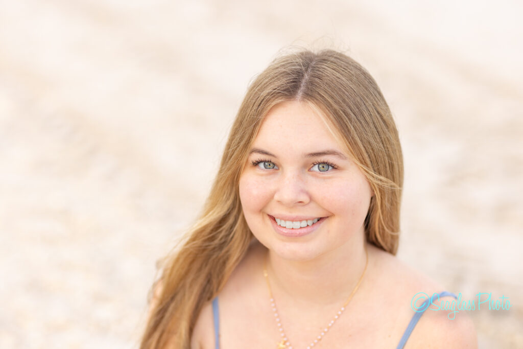 A young woman with long blonde hair smiles at the camera. She is outdoors with a blurred, sandy background and is wearing a necklace and a sleeveless top. Vero Beach Photoshoot