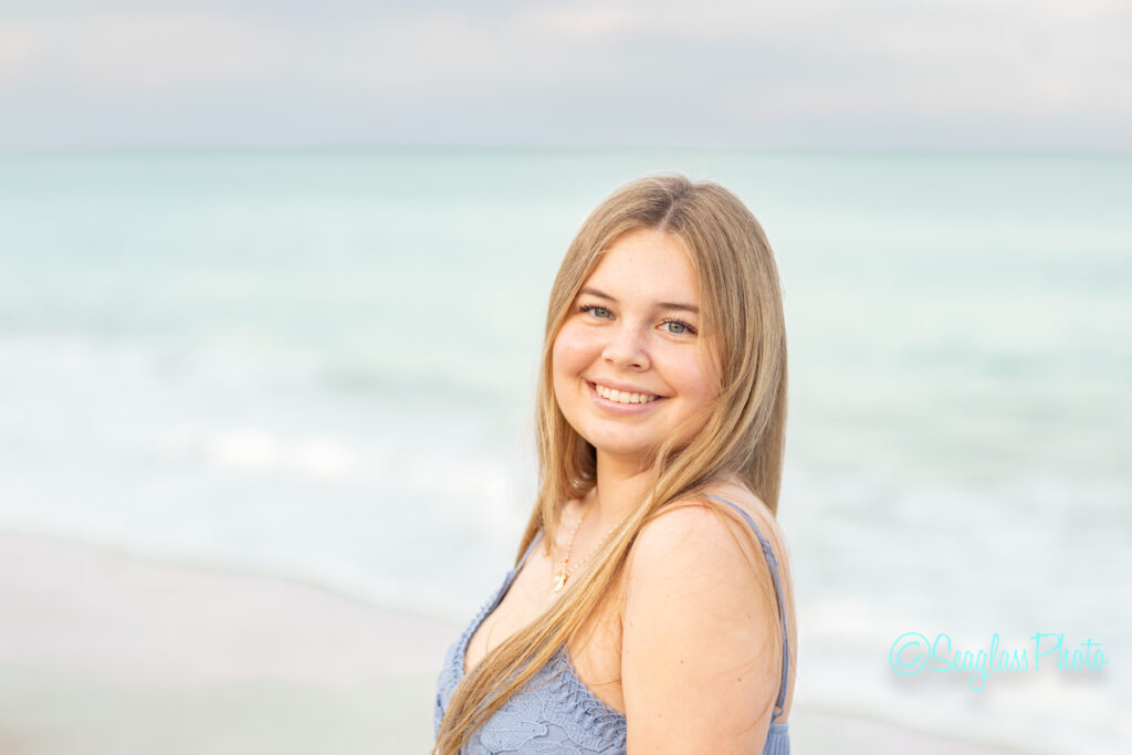 A young woman with long blonde hair smiles at the camera while standing on a beach with light blue ocean waves and a cloudy sky in the background. Vero Beach Photoshoot