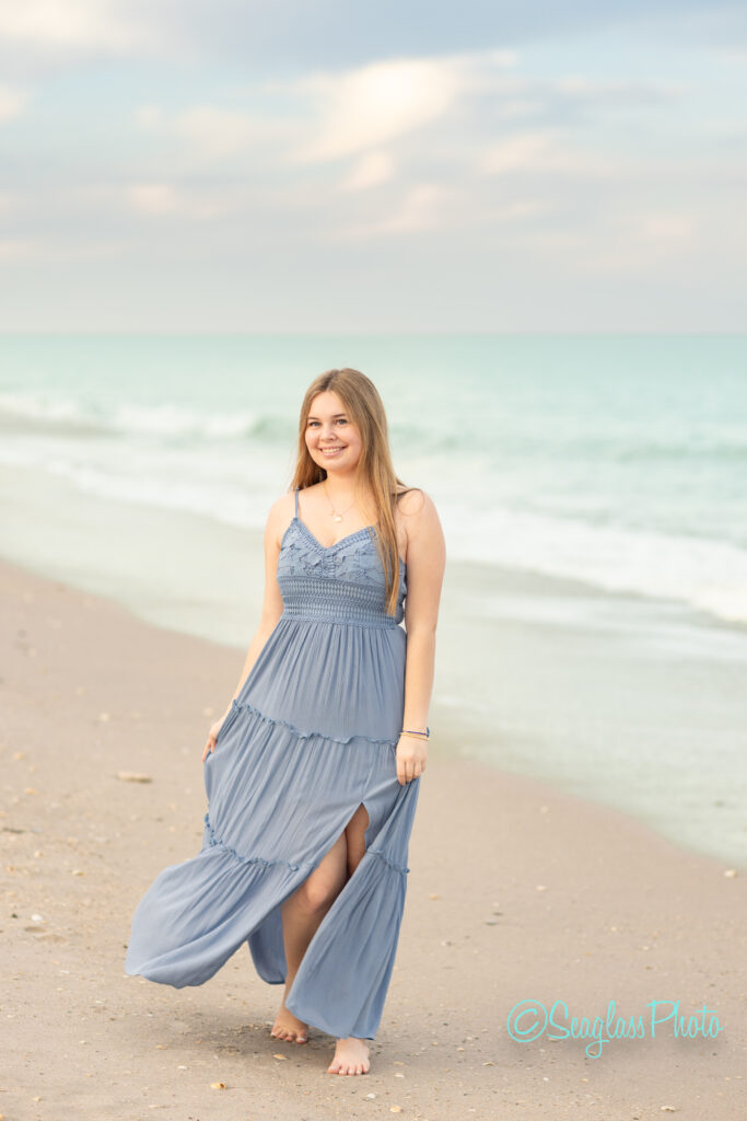 A young woman in a flowing blue dress walks barefoot along a sandy beach near the ocean, smiling with the sea and sky softly blurred in the background. Vero Beach Photoshoot