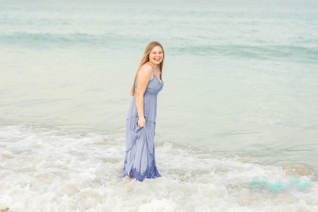 A young woman in a long, light purple dress stands smiling in shallow ocean water, with gentle waves around her feet and the sea in the background. Vero Beach Photoshoot
