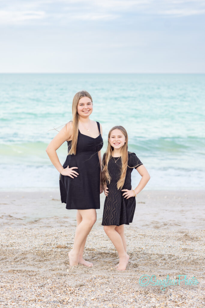 Two girls with long blonde hair, both wearing black dresses, stand barefoot on a sandy beach with the ocean and a cloudy sky in the background. They are smiling and have their hands on their hips. Vero Beach Photoshoot