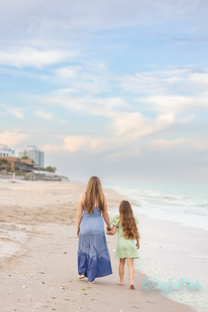 A woman and a young girl with long hair walk hand-in-hand along a sandy beach, facing the ocean. The sky is partly cloudy, and waves gently lap the shore. Buildings are visible in the distance. Vero Beach Photoshoot