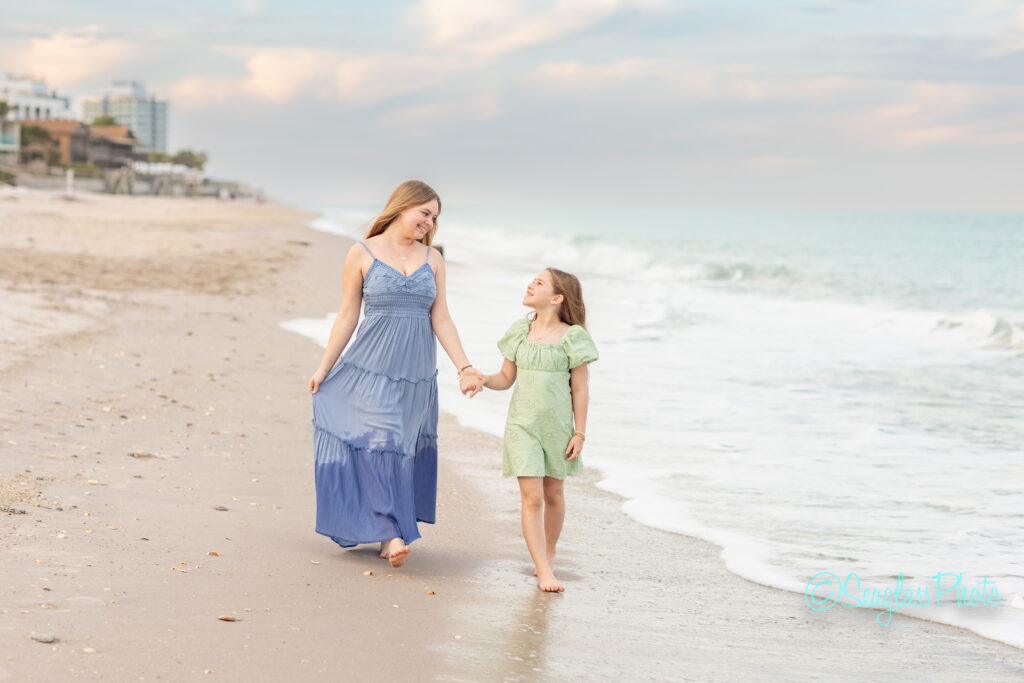 A woman in a long blue dress and a young girl in a green dress walk barefoot, holding hands and smiling at each other, along the shoreline of a sandy beach with gentle waves. Vero Beach Photoshoot