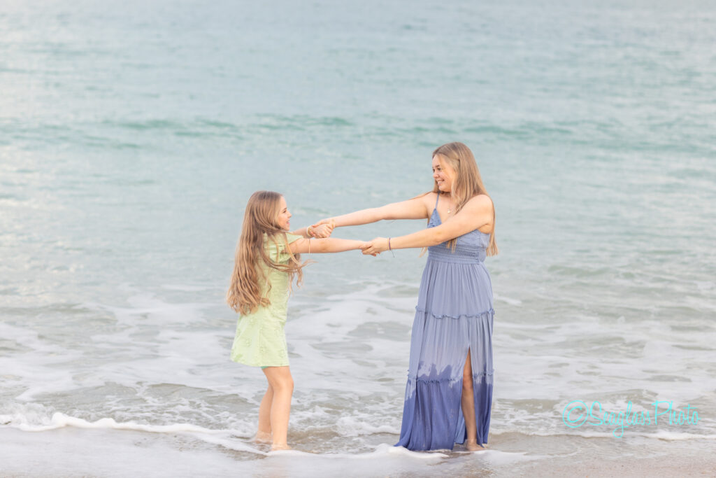 A woman in a blue dress and a young girl in a light green dress hold hands and smile at each other while standing in shallow ocean water at the beach. Vero Beach Photoshoot