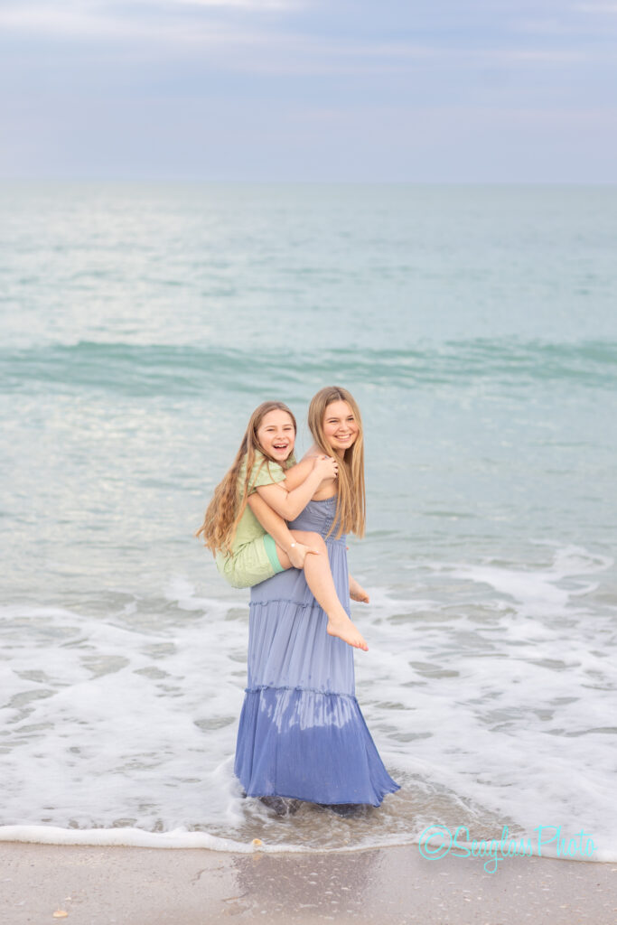 Two young girls smile joyfully at the beach, one wearing a light green dress and being carried piggyback by the other in a long blue dress, with gentle waves and a calm sea in the background. Vero Beach Photoshoot