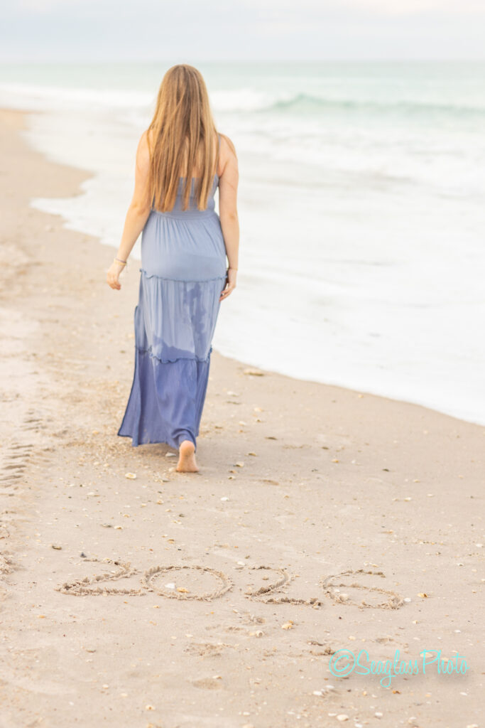 A woman in a blue dress walks barefoot along the beach near the ocean, with “2026” written in the sand ahead of her. Vero Beach Photoshoot