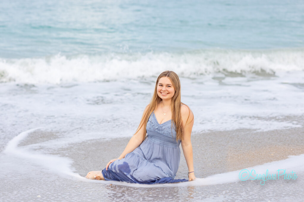 A young woman in a light blue dress sits on the wet sand at the edge of the beach, smiling at the camera as gentle waves wash in behind her. Vero Beach Photoshoot