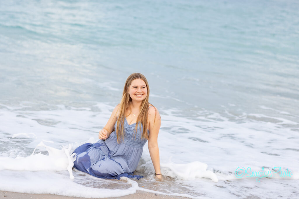 A young woman in a light blue dress sits on the shore where gentle ocean waves wash over her legs, smiling at the camera under a cloudy sky. Vero Beach Photoshoot