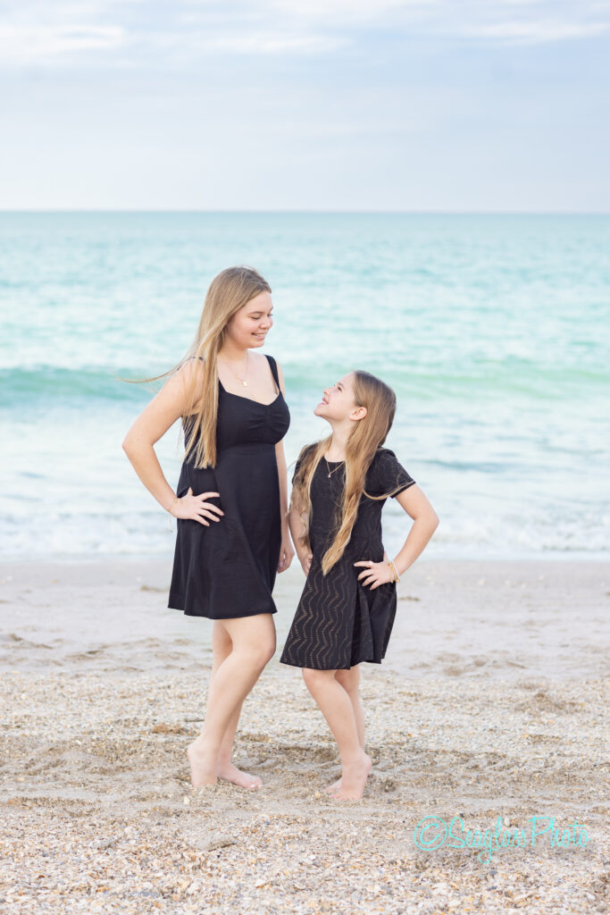 Two girls with long blonde hair, wearing black dresses, stand barefoot on a sandy beach. They face each other and smile, with the ocean and cloudy sky in the background. Vero Beach Photoshoot