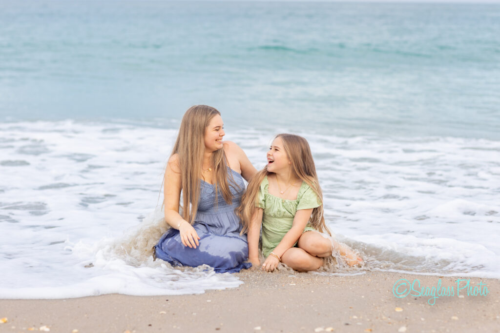 A woman and a young girl sit on the beach at the edge of the ocean, smiling and laughing together as gentle waves wash over their legs. The woman wears a blue dress, and the girl wears a light green dress. Vero Beach Photoshoot