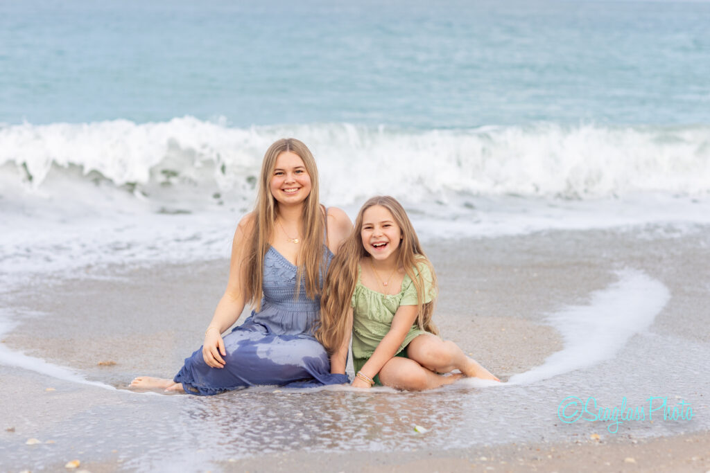 Two girls with long hair sit on wet sand at the beach, smiling at the camera. Waves and ocean water rush up behind them. They wear light summer dresses, one blue and one green. Vero Beach Photoshoot