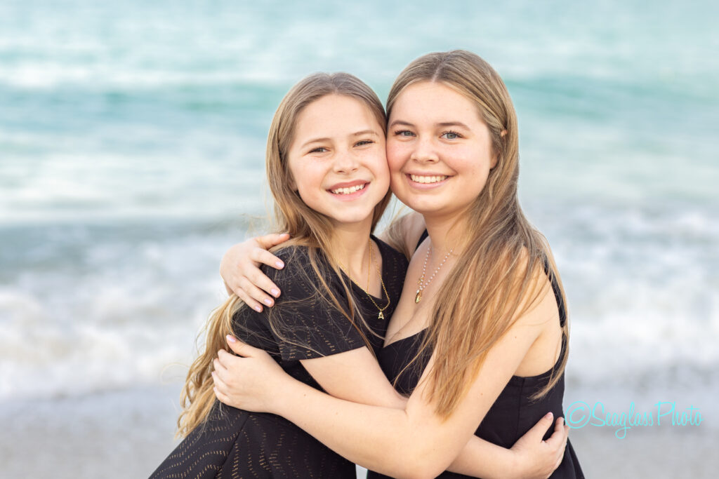 Two smiling young women with long hair, wearing black dresses, stand on a beach hugging each other. The ocean waves and sandy shore are visible in the background. Vero Beach Photoshoot