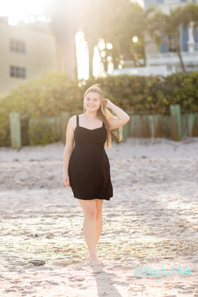 A young woman with long hair, wearing a sleeveless black dress, stands barefoot on a sandy beach, smiling at the camera with one hand touching her hair. The sun shines brightly in the background. Vero Beach Photoshoot