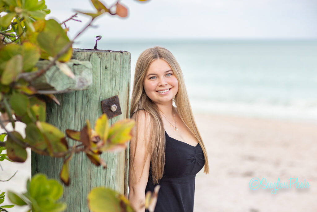 A young woman with long blonde hair wearing a black dress leans against a weathered wooden post on a sandy beach, smiling at the camera. The ocean and some plant leaves are visible in the background. Vero Beach Photoshoot