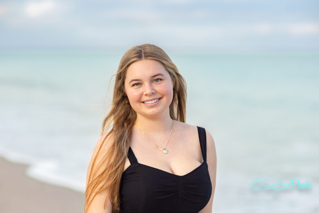 A young woman with long blonde hair, wearing a black sleeveless dress and a pendant necklace, smiles while standing on a sandy beach with calm, turquoise water in the background. Vero Beach Photoshoot