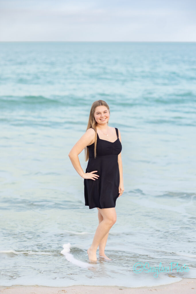 A young woman in a black dress stands barefoot at the edge of the ocean, smiling at the camera. The water is calm and the sky is clear, creating a serene beach scene. Vero Beach Photoshoot