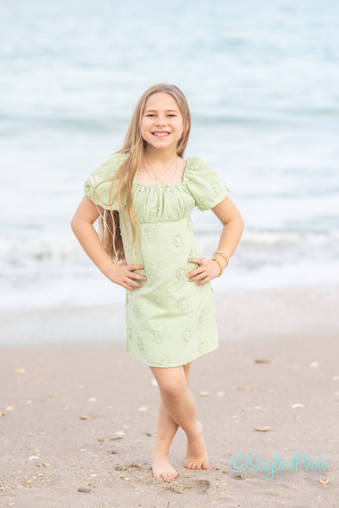 A smiling young girl with long blonde hair stands barefoot on a sandy beach, wearing a light green dress. The ocean waves are visible in the background. Vero Beach Photoshoot