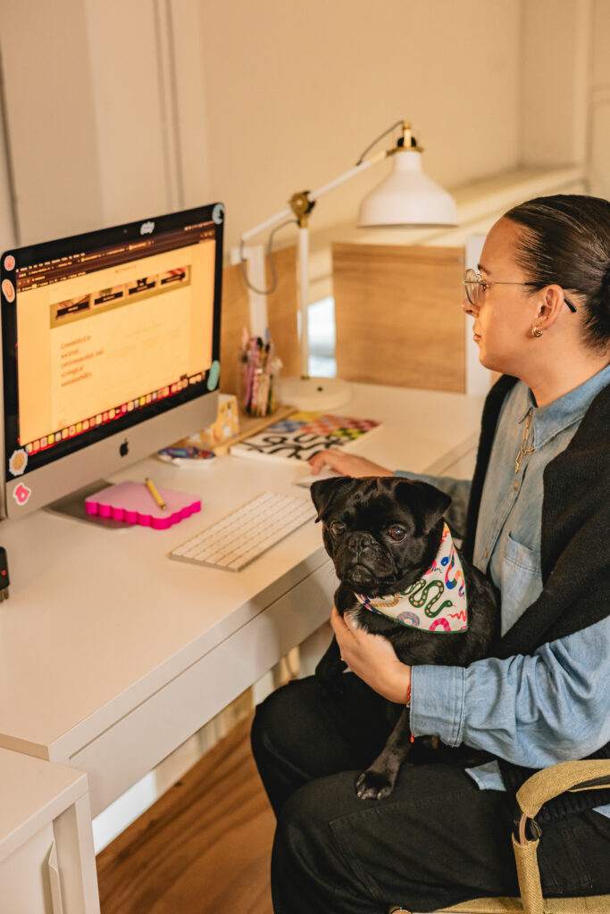 A person sitting at a desk works on a computer while holding a black pug wearing a colorful bandana. The workspace includes a lamp, keyboard, and desk accessories. Vero Beach Photoshoot