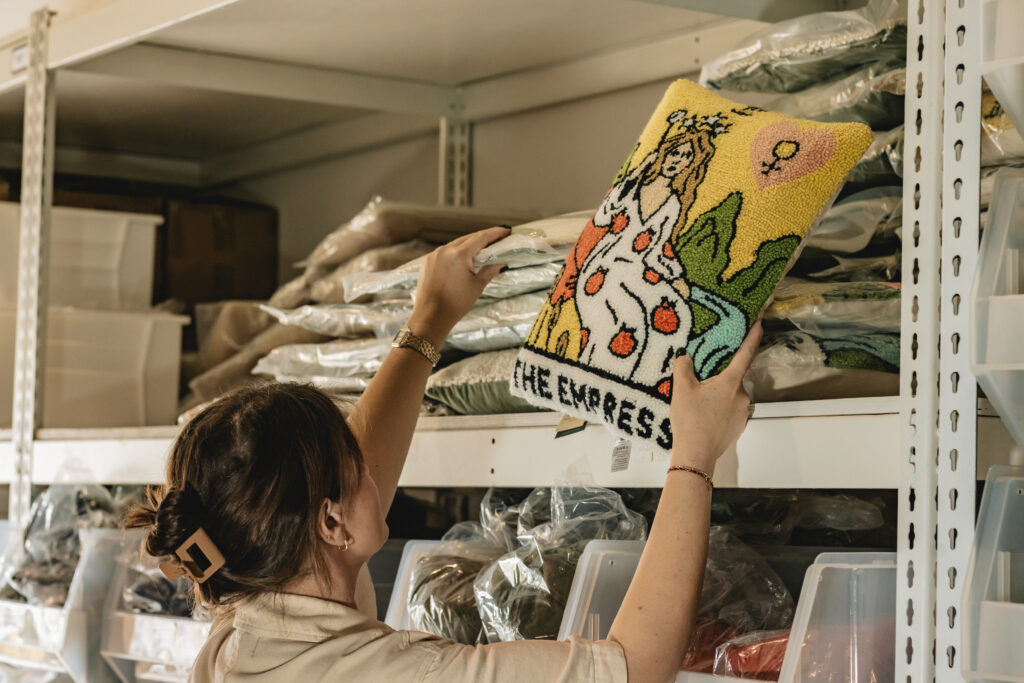 A person places a colorful, embroidered cushion labeled THE EMPRESS depicting a woman in a crown on a shelf filled with stacked fabric and plastic bins in a storage area. Vero Beach Photoshoot