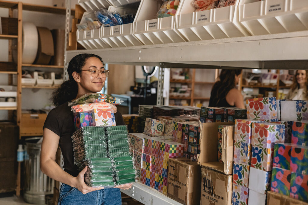 A woman wearing glasses holds a large stack of colorful packaged items while standing by shelves filled with neatly organized products in a storage room. Vero Beach Photoshoot