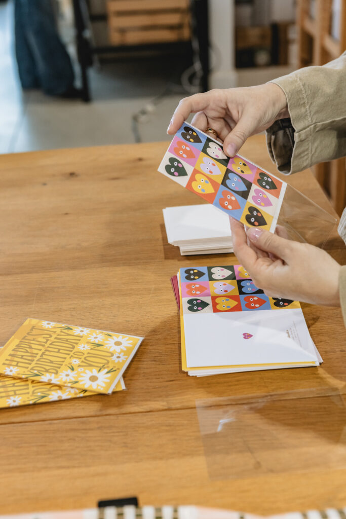 A person holds a colorful greeting card with a pattern of cartoon owl faces over a wooden table, with other cards and envelopes scattered nearby. Vero Beach Photoshoot