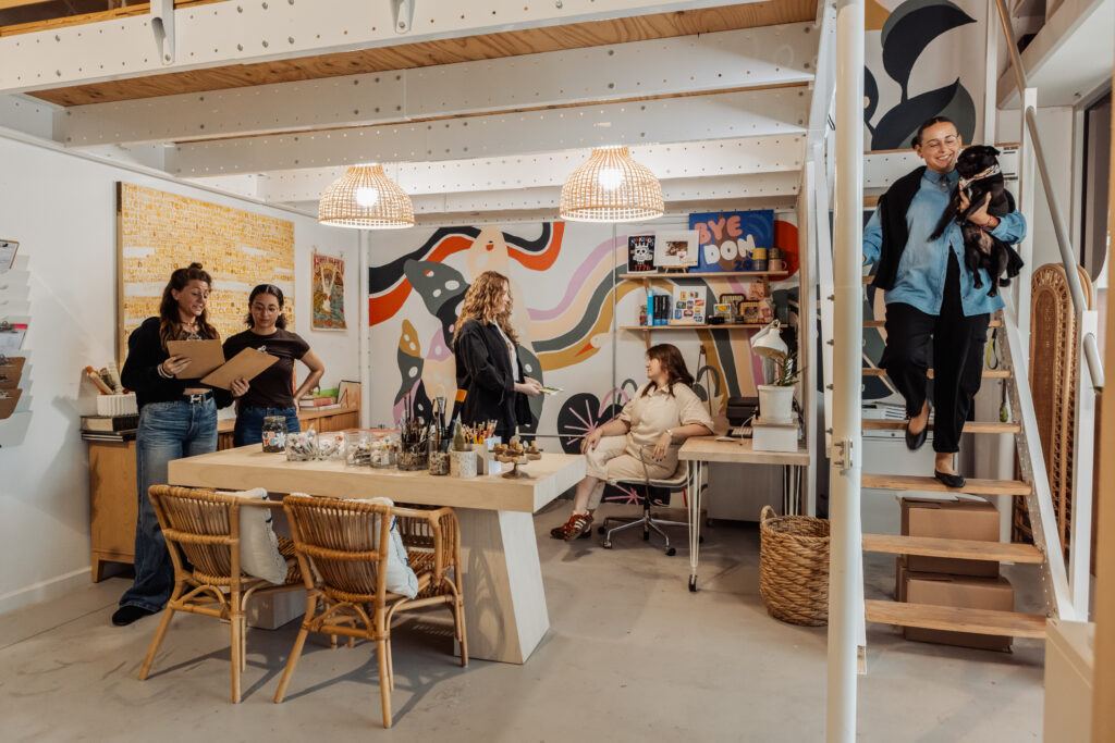 Five women are in a colorful, modern office studio. Three stand together chatting, one sits at a desk, and another walks down stairs holding a small dog. Art supplies and decorations fill the bright, creative space. Vero Beach Photoshoot
