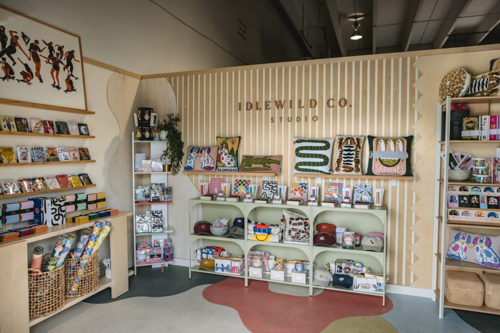 A boutique shop interior with wooden panel walls displays colorful gift items, greeting cards, plush pillows, mugs, books, and baskets on shelves. Art prints and plants decorate the cozy, well-lit space. Vero Beach Photoshoot