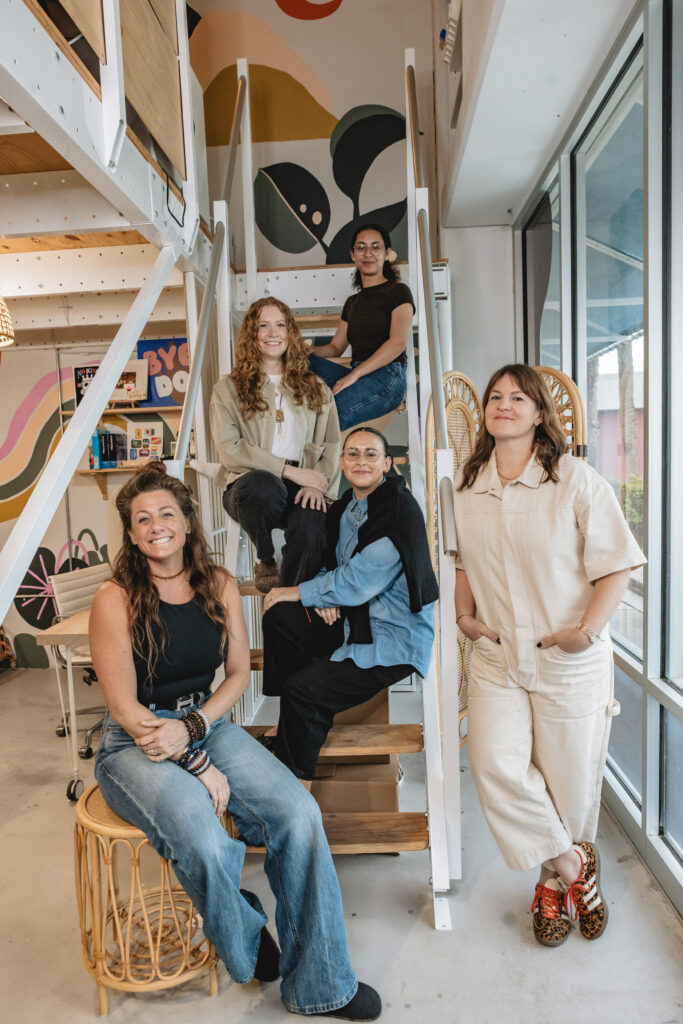 Five women pose together on and around a staircase in a modern, colorful office space with large windows and murals on the walls. They are smiling and casually dressed, creating a friendly, relaxed atmosphere. Vero Beach Photoshoot