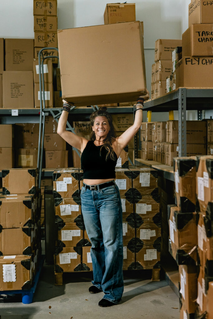 A smiling woman in a black tank top and jeans stands in a warehouse, lifting a large cardboard box over her head. She is surrounded by shelves and stacks of brown boxes. Vero Beach Photoshoot