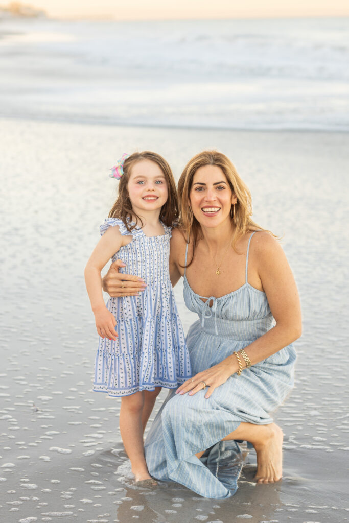 A woman kneels beside a young girl on a sandy beach near the water’s edge. Both are smiling and wearing light blue summer dresses. The beach and ocean waves are visible in the background. Vero Beach Photoshoot