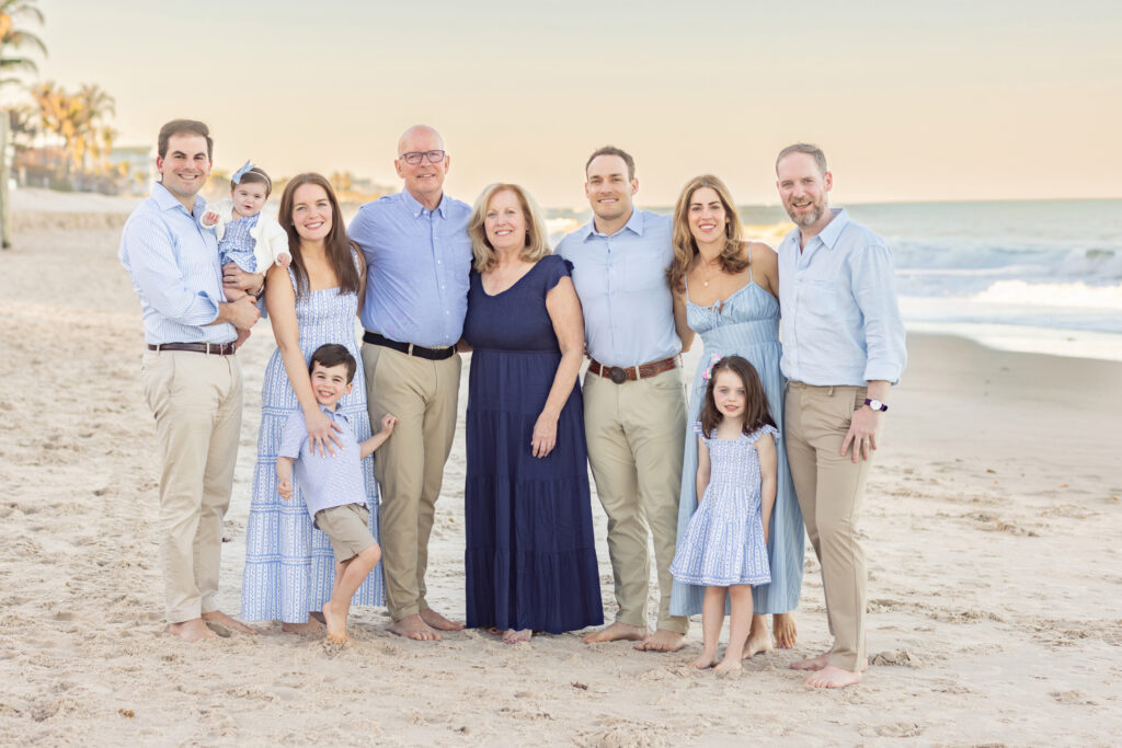 A family of ten poses barefoot on a sandy beach at sunset. Adults and children, dressed in coordinated blue and white outfits, stand together smiling with the ocean and palm trees in the background. Vero Beach Photoshoot