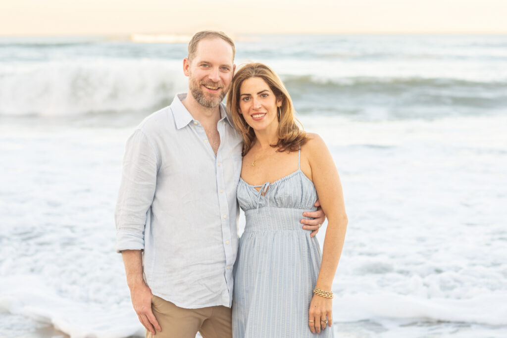 A man and woman stand close together on a beach, smiling at the camera. The woman has her arm around the man. Gentle ocean waves and a clear sky are visible in the background. Vero Beach Photoshoot