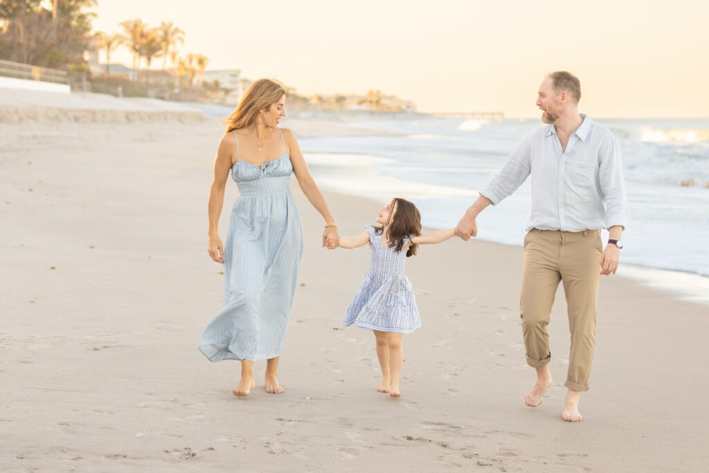 A woman, a man, and a young girl hold hands and walk barefoot along the beach, smiling at each other with the ocean and palm trees in the background. Vero Beach Photoshoot