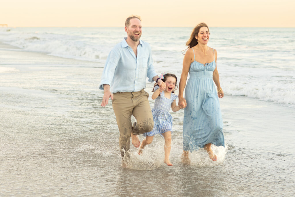 A smiling man, woman, and young girl run barefoot on a beach, holding hands and splashing in the shallow waves with the ocean and soft orange sky in the background. Vero Beach Photoshoot