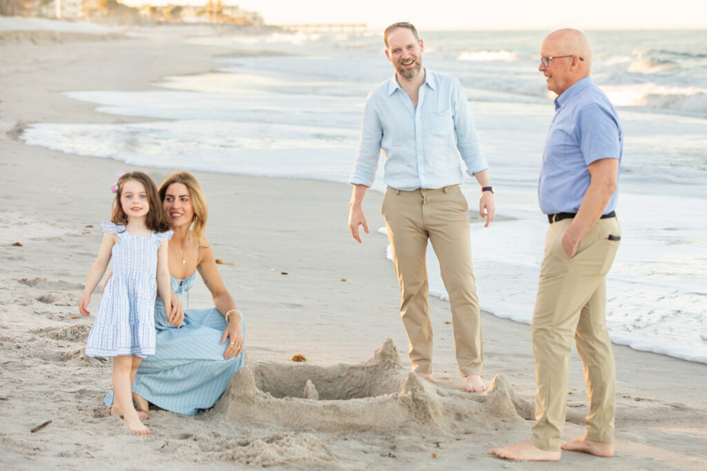 A young girl, a woman, and two men smile by a sandcastle on a beach. The woman kneels beside the girl, while the men stand nearby. Ocean waves and a distant pier are visible in the background. Vero Beach Photoshoot